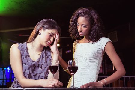 Zwei Frauen mit Weingläsern, eine fasst sich an den Kopf, die andere unterstützt sie.