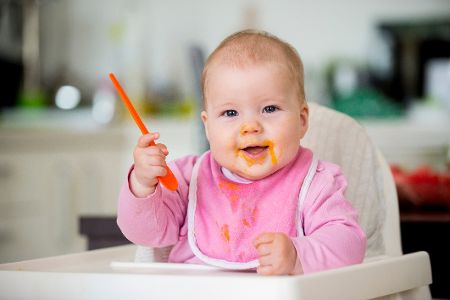 Baby sitzt in Hochstuhl, hat beschmierten Mund und einen Löffel in der Hand Baby sitzt in Hochstuhl, hat beschmierten Mund und einen Löffel in der Hand