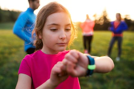 Kind steht auf einer Wiese neben anderen Menschen und schaut auf seine Smartwatch