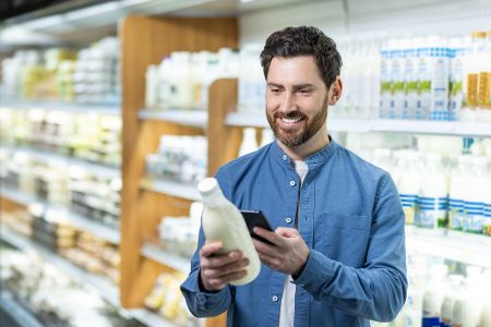 Lächelnder Mann im Lebensmittelgeschäft mit Smartphone zur Auswahl von Milchprodukten aus dem Kühlregal Lächelnder Mann im Lebensmittelgeschäft mit Smartphone zur Auswahl von Milchprodukten aus dem Kühlregal