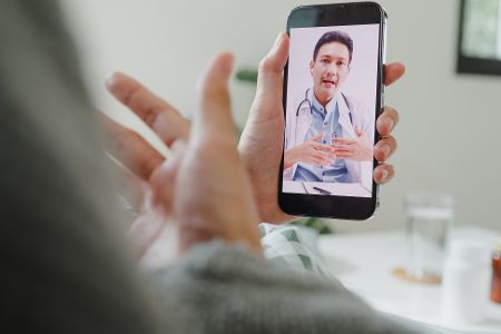 Jemand hält ein Handy in der Hand, auf dessen Bildschirm eine Arzt im Videogespräch zu sehen ist.
