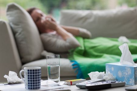 Im Hintergrund liegt eine kranke Frau auf dem Sofa und putzt sich die Nase. Vorne auf dem Tisch stehen Taschentücher und Medikamente. Im Hintergrund liegt eine kranke Frau auf dem Sofa und putzt sich die Nase. Vorne auf dem Tisch stehen Taschentücher und Medikamente.