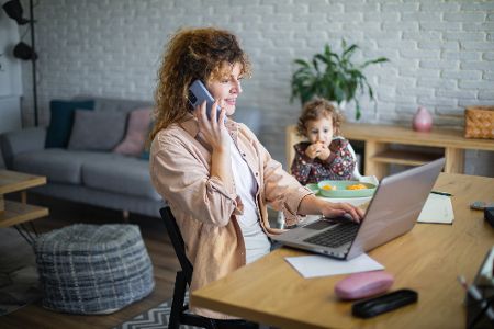 Frau sitzt im Homeoffice vor dem Rechner und telefoniert, neben ihr ein Kleinkind im Hochstuhl.