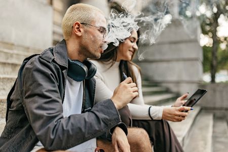 Zwei junge Menschen sitzen draußen auf einer Treppe und rauchen E-Zigaretten. Zwei junge Menschen sitzen draußen auf einer Treppe und rauchen E-Zigaretten.