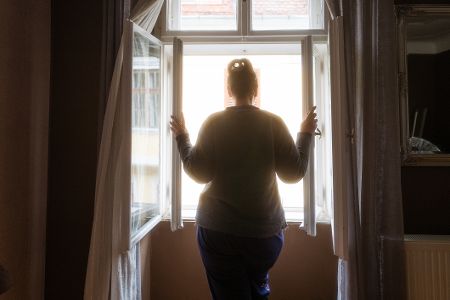 Frau steht vor dem geöffneten Fenster in einem Zimmer und schaut nach draußen