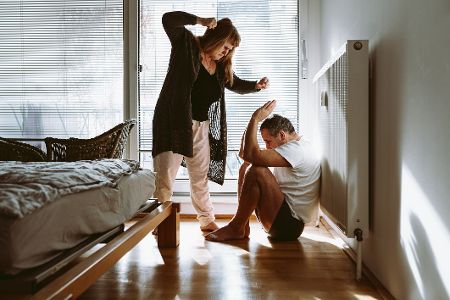Mann sitzt im Schlafzimmer auf dem Fußboden, hebt abwehrend die Hände gegenüber einer Frau, die vor ihm steht und ihn schlagen will