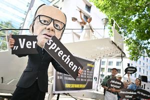 Ein Aktivist mit Merz-Maske zerbricht während einer symbolischen Protestaktion von Kabul Luftbrücke und Campact ein Schild mit der Aufschrift "Versprechen". 