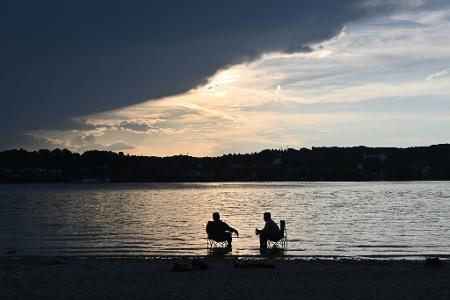 Dunkle Wolken ziehen über den Starnberger See während zwei Männer mit Campingstühlen im Wasser sitzen.